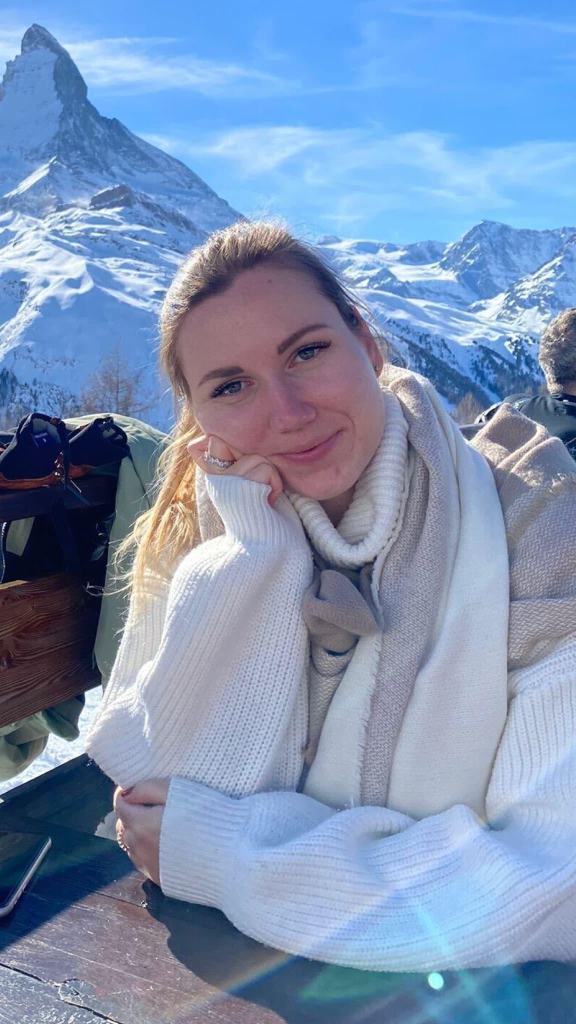A woman is sitting outside and resting her cheek on her closed hand with a snowy mountain peak in the background