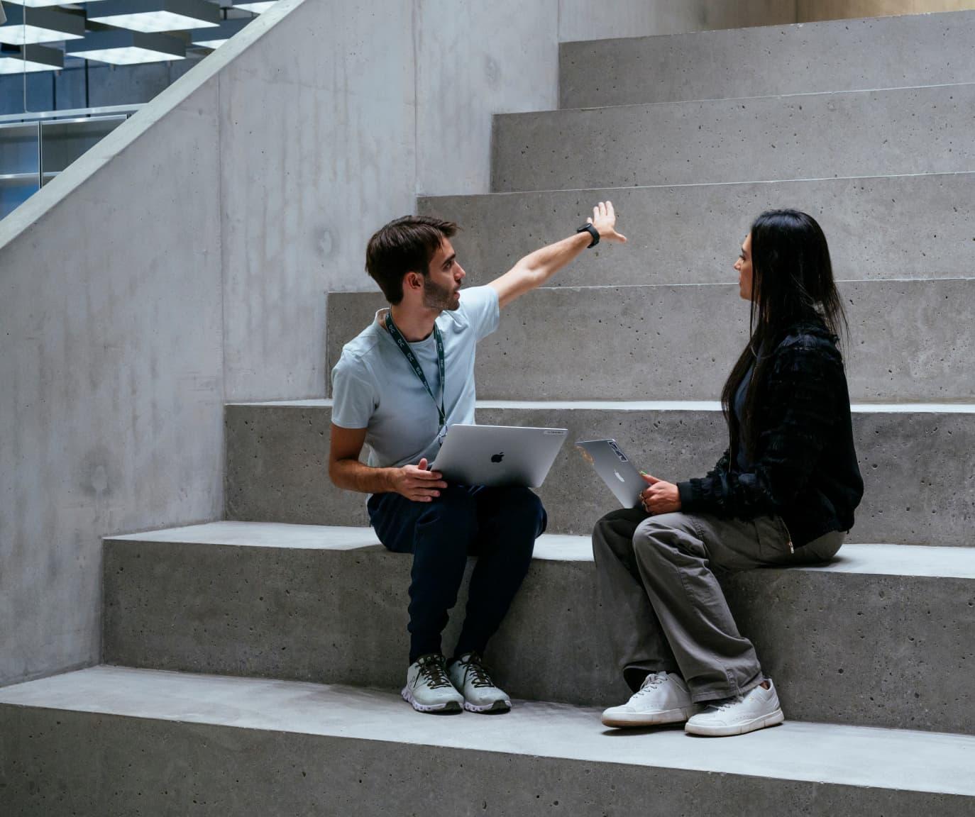 Woman and man sat on concrete steps looking at each other as the man is pointing towards the top of the steps